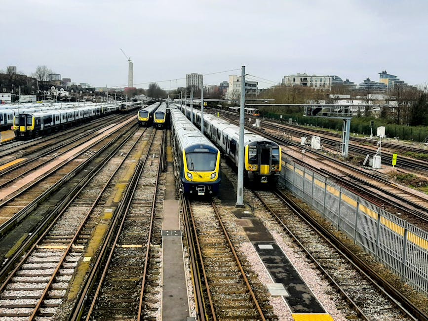 A view of multiple modern commuter trains parked on parallel railway tracks at a train station near Finsbury Park. The trains have sleek, yellow and grey external finishes with large windows. The platforms are visible on the sides, with some areas appearing clean and well-maintained. Overhead electric lines and poles run above the tracks, and beyond, there are urban buildings and trees under a cloudy sky. The scene captures the infrastructure involved in surface cleaning and maintenance for efficient transportation services, reflecting the importance of thorough, professional cleaning and sanitisation for public transit environments, as provided by Cleaner Finsbury Park.