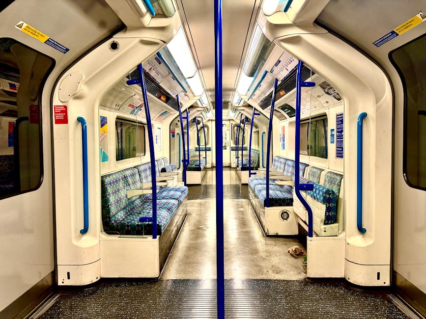 Inside a clean, well-maintained train carriage with a symmetrical layout, featuring blue and purple patterned fabric seats arranged in rows along both sides. The surface of the seats appears freshly cleaned and free of dust or stains, with the upholstery looking smooth and tidy. The beige plastic walls and ceiling are spotless, reflecting the bright overhead lighting, which enhances the cleanliness and hygienic appearance of the space. Blue handrails and vertical bars are securely fixed, free of smudges or grime. The floor is covered with a dark, non-slip rubber surface that remains dry and clean, with minimal debris visible, indicating recent thorough cleaning by Cleaner Finsbury Park as part of their domestic cleaning and sanitisation services. Large windows along both sides reveal the station environment outside, contributing to the bright and airy atmosphere of the carriage, emphasizing the importance of surface cleaning and hygiene maintenance in public transport settings.