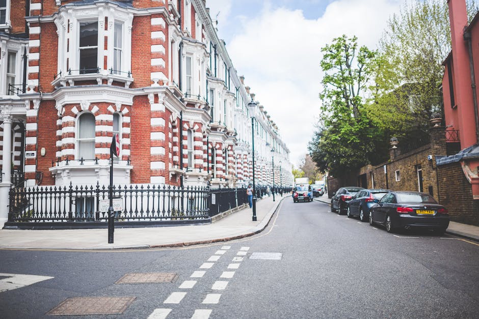 A residential street in Finsbury Park, featuring a row of historic red-brick Victorian buildings with white decorative details, bay windows, and black wrought-iron railings. The street is lined with parked cars including sedans and hatchbacks, and has a clear, paved road with white dashed lines and pedestrian crossing markings. Green trees with fresh leaves canopy the area, and a lamppost stands near the corner. Bright daylight illuminates the scene, highlighting the cleanliness and well-maintained appearance of the neighborhood, as potentially cleaned and maintained by Cleaner Finsbury Park in alignment with residential and surface cleaning services.