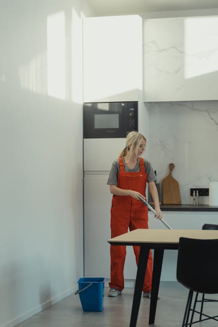 Two women are engaging in domestic cleaning inside a modern living room with a light grey wall adorned with black and white framed artwork. One woman with curly hair, wearing a beige shirt and black t